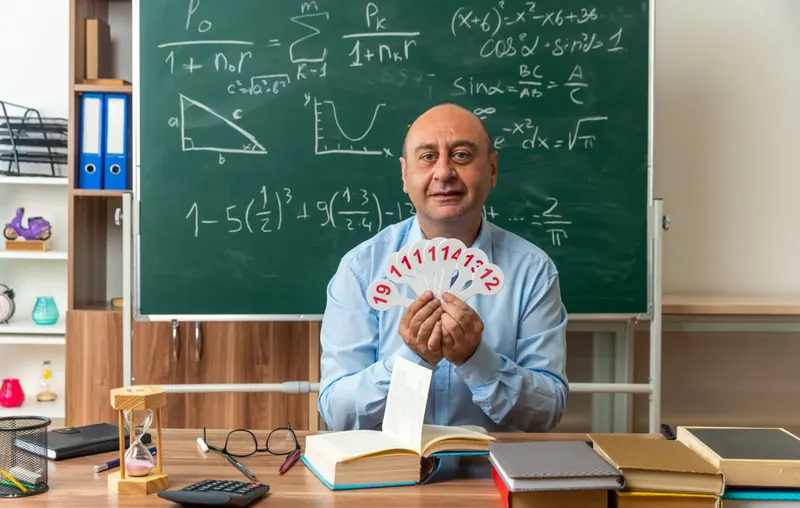 معلّم على مكتبه أمام السبورة، يحمل بطاقات أرقام